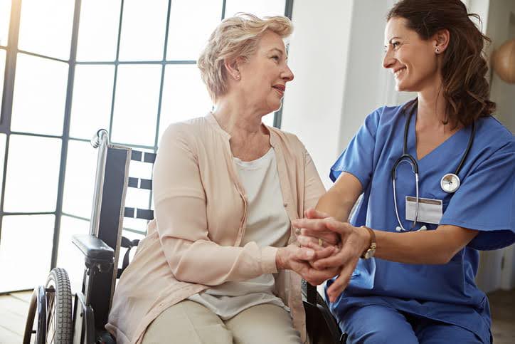 elderly woman in wheelchair speaking to female nurse in blue scrubs who is smiling. 