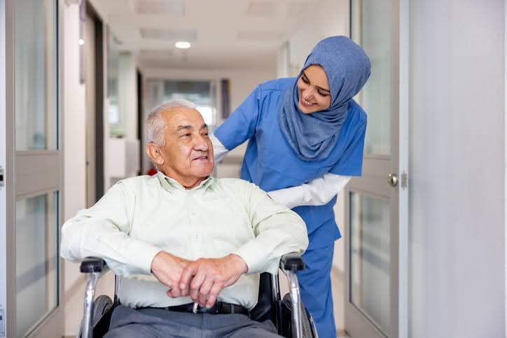 A nurse in a skilled nursing facility in Salt Lake City pushing an elderly man in a a wheelchair down the hall while they smile and talk to one another