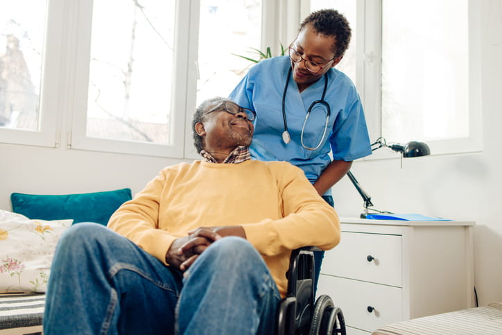 A nurse and patient smile at one another as she assists him in his wheelchair at a skilled nursing facility. 