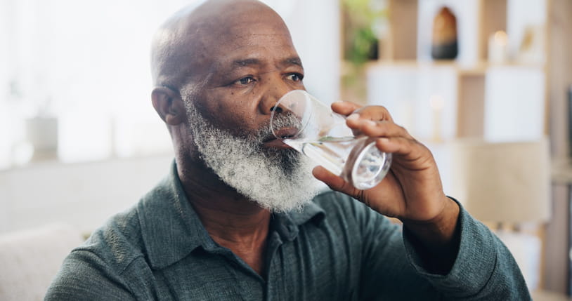An older African American man, bald with a gray beard, drinks a glass of water while sitting indoors in a warmly lit room