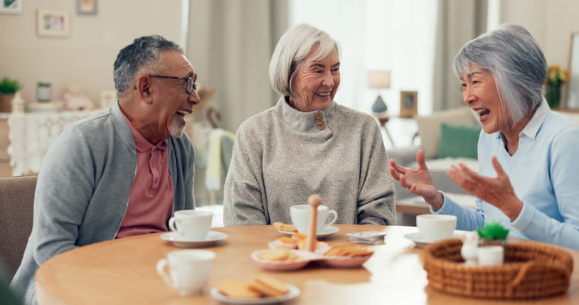 A diverse group of three happy seniors—one man and two women—are laughing together while enjoying a conversation over coffee and snacks at a round wooden table.