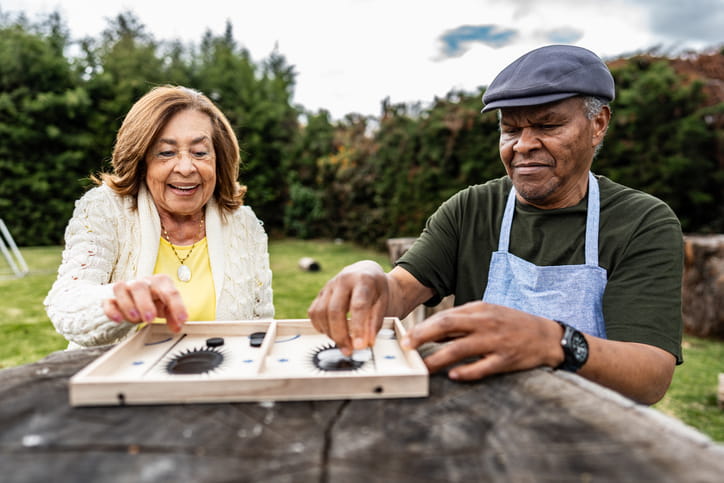  A diverse senior couple, a Hispanic woman and a Black man, are sitting at a wooden table outdoors and smiling as they play a board game together.