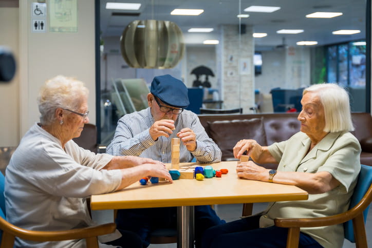 Three elderly people—a man and two women—are sitting at a table in a community room, focused on playing a wooden block-stacking game together.