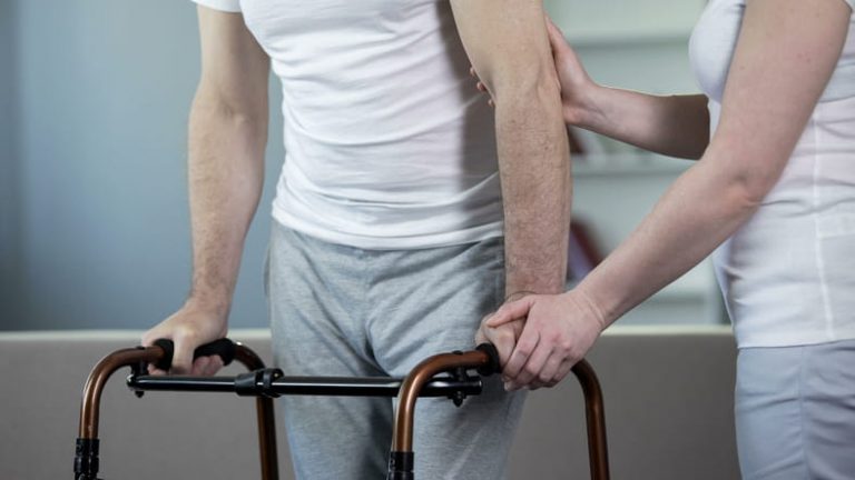 A female caregiver in a white uniform provides support to a male patient who is using a walker. The close-up shot focuses on her hands steadying his arm as he holds the walker's grips.