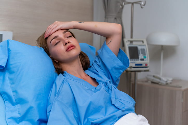 A young woman in a blue hospital gown lies in a hospital bed with her eyes closed and a hand on her forehead, appearing to be in pain or feeling unwell. An IV stand is visible in the background.