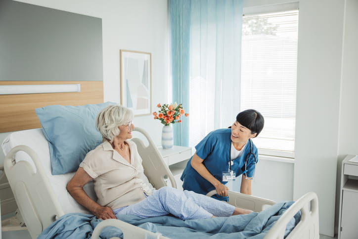 In a bright hospital room, a female nurse in scrubs smiles while adjusting the bed for a senior woman. The patient is sitting up comfortably and smiling back at the nurse.