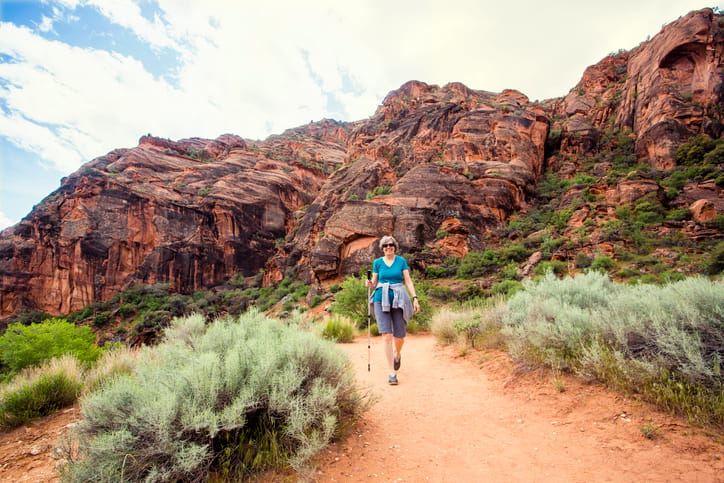 An active senior woman with sunglasses and a trekking pole smiles as she hikes on a dirt trail through a desert canyon with large red rock formations in the background.