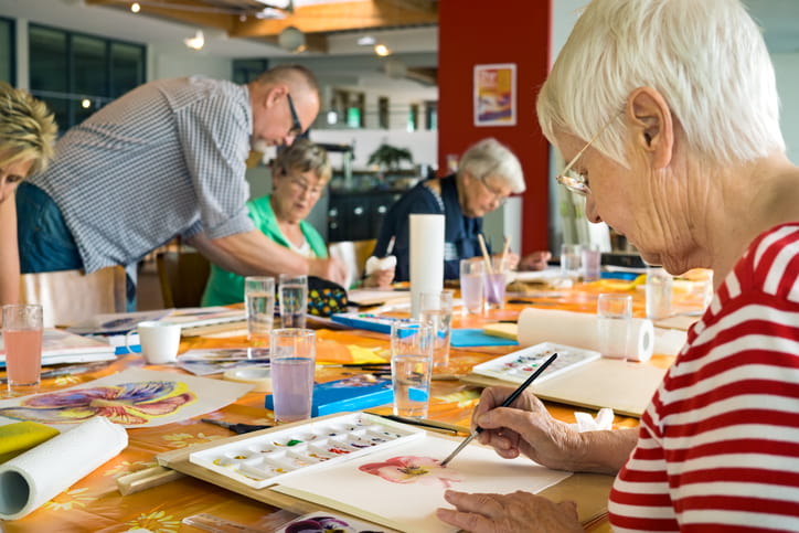 A group of seniors participates in a watercolor painting class in a bright room. The foreground focuses on a woman with white hair painting a flower, while other students and an instructor are at a long table in the background.
