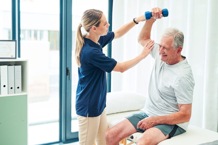 A female physical therapist helps a senior man lift a light blue dumbbell over his head while he sits on an examination table, focusing on arm strengthening exercises