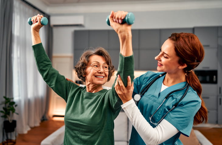 A smiling senior woman lifts light dumbbells as a female nurse in scrubs supports her arm during a positive and encouraging in-home physical therapy session