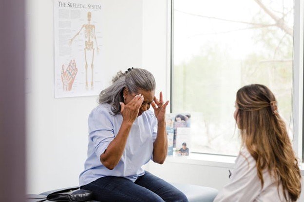 An elderly woman holding her head as she speaks with her doctor.