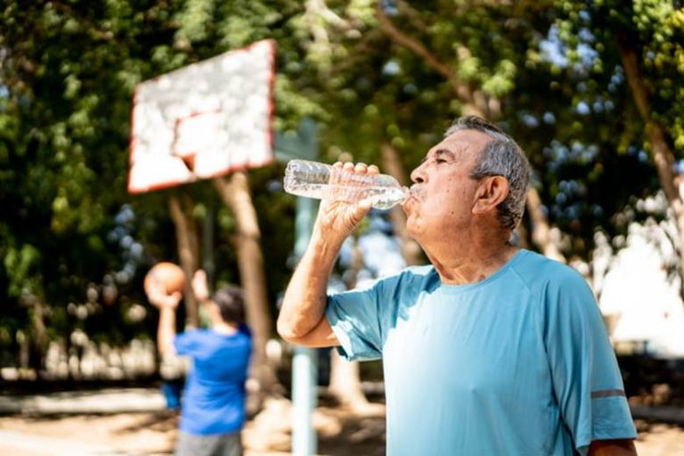 An elderly man drinks water while outside in the summer heat.