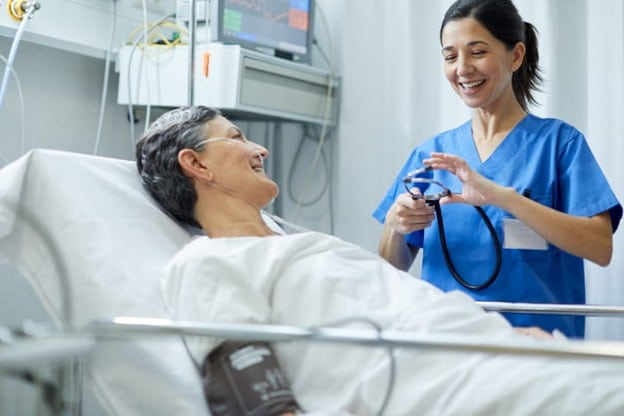 Understanding Hospital Deconditioning And The Rehabilitation Process 2 A female nurse laughs with a patient who is in a hospital bed.